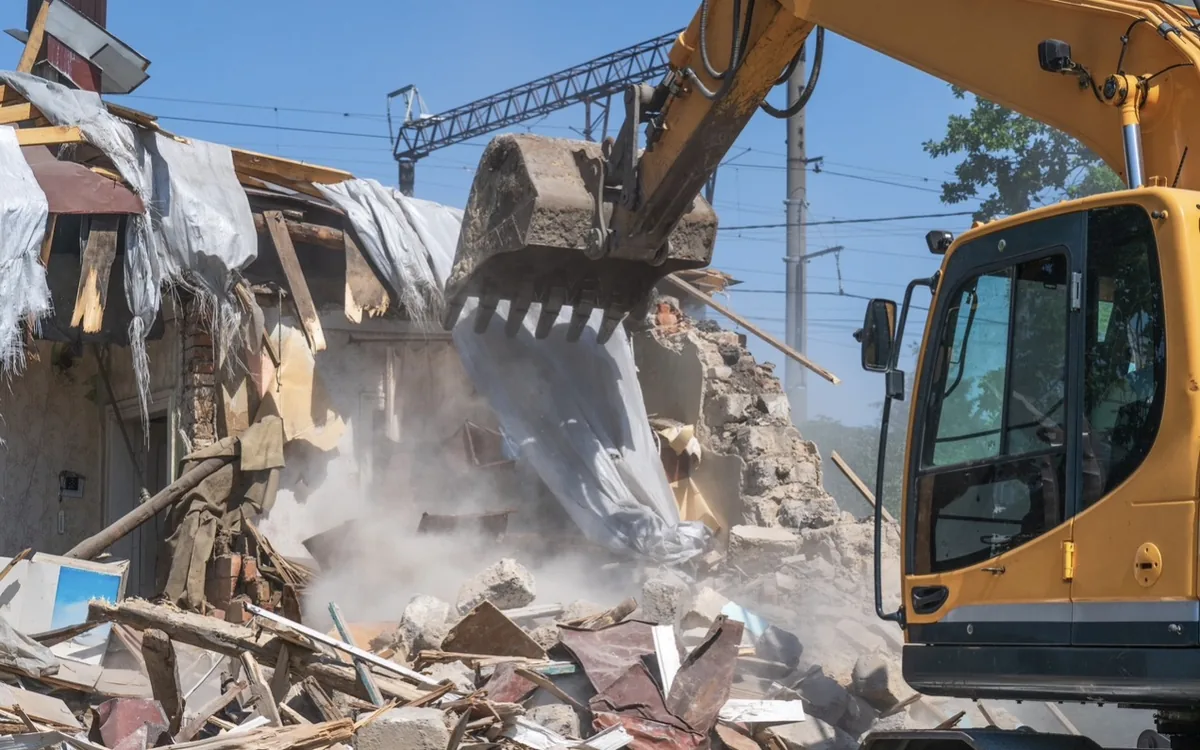 Excavator tearing down a brick building with dust rising from the rubble